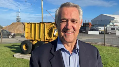 A man with grey hair and a smart jacket smiles in the sunshine. Behind him is an old mine excavator and in the distance the scaffold minehead and wheel of South Crofty Mine. A Cornish flag flutters on the top  