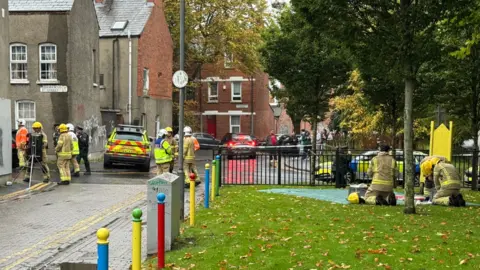 A number of firefighters and police officers are standing around the bottom of Damascus Street. There are police cars parked along the bottom of the street and some cordon tape placed.