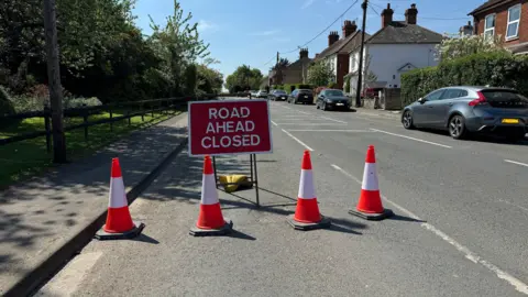 BBC Badshot Lea Road near Farnham. A red sign says 'ROAD AHEAD CLOSED' in white letters. There are four red and white traffic cones in front of it. There are trees and plants on one side of the road, on the other are houses and cars.