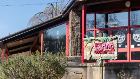 Wild Planet The picture shows the exterior of a building labelled "Island Restaurant." The structure features large windows with red frames and a stone facade. In front of the building, there are bushes and trees, adding greenery to the scene. The restaurant sign is decorated with tropical-themed elements like leaves.
