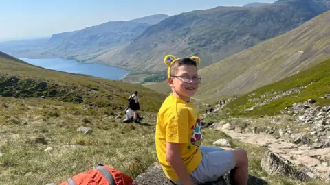 Family photo A boy with brown hair and glasses in a yellow T-shirt and white shorts sat on a rock, part way up a large hill which can be seen stretching down to a lake below and there are other large hills in the distance