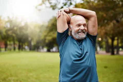 Getty Images Man stretching with arms above head in a park with green grass. The man has a grey beard, is bald and wears a blue t-shirt. He is smiling. 