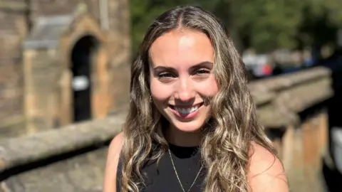 BBC Orla Sonvico smiling at the camera. She has long brown hair and is wearing a black top. There is a wall and an old sandstone building in the distance.