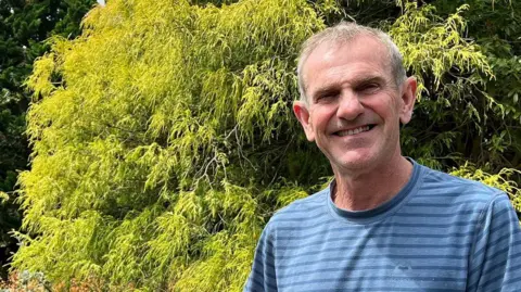 BBC Gregory Quinn stands in his front garden with a thick green hedge behind him. He is wearing a blue striped t shirt and is smiling at the camera. 