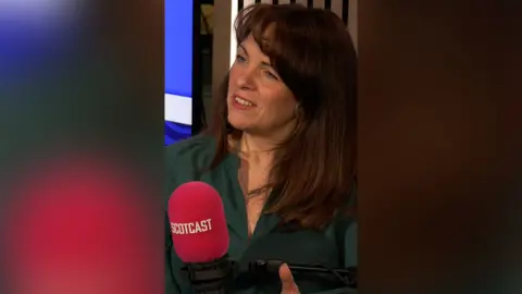 Catriona MacPhee in a green shirt speaking into a pink microphone with the word 'Scotcast' on it.