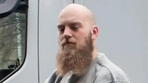 Athena Alexander Dighton, he is bald with a light brown beard. He is wearing a grey sweatshirt. His eyes are closed.