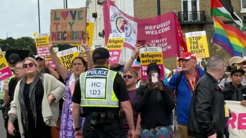 BBC A group of protesters hold up banners saying Stop the Far Right and Refugees Welcome. A police officer stands in front of the group with his back to the camera.