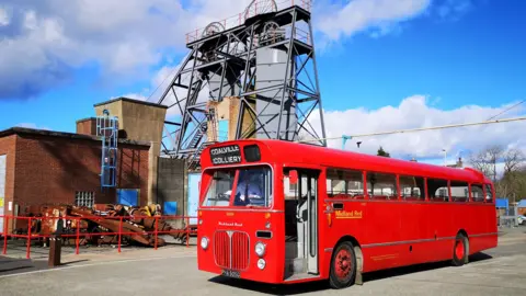 Leicester Transport Heritage Trust A vintage red single-decker bus in front of a mining colliery wheel.