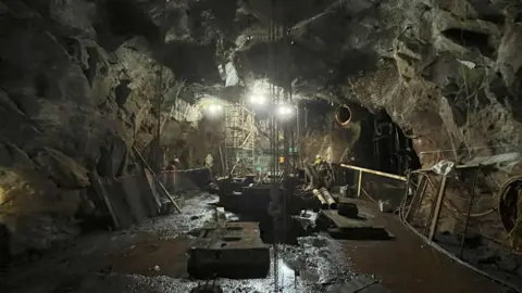 Cornish Metals A dark cavern below ground all around is heavy equipment, chains and scaffolding. Floodlights illuminate the scene. 