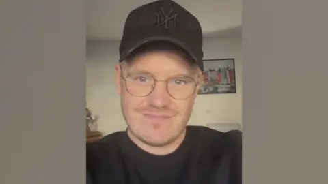 A selfie of Ryan Forey. He is wearing a black cap and top, and has rounded glasses. He is standing in front of a picture of a row of buildings. 