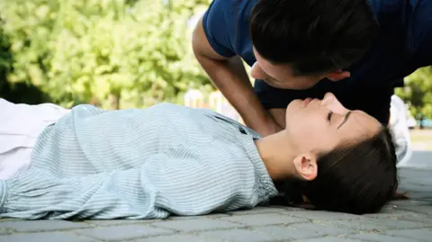 Getty Images A man listens to see if a woman is breathing. She is lying on the floor wearing a blue stripped shirt and white trousers. She has long dark hair. The man is bending over her wearing a blue tshirt.