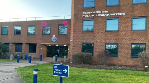 The front of Bedfordshire Police headquarters. It is a red brick building with grass and bushes outside. In the foreground there is a sign for visitors parking.