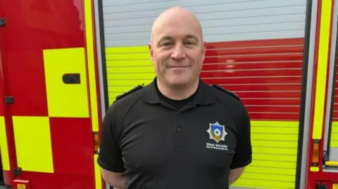 West Yorkshire Fire and Rescue Service West Yorkshire Fire and Rescue Services' Assistant District Commander David Burland. wears a black polo shirt embroidered with the force's logo and stand in front of a fire engine. 