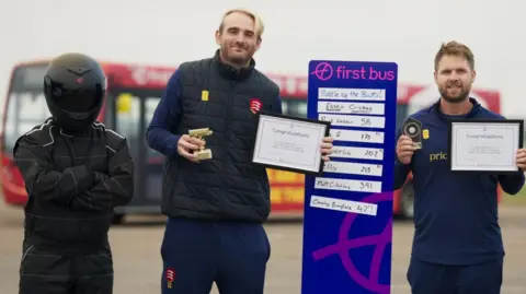 Matt Critchley, Paul Walter and First Bus' Stig, who is wearing all black and a black helmet, standing next to a purple leaderboard. Critchley and Walter are holding certificates, while Walter also holds a gold trophy. A red bus is behind them.