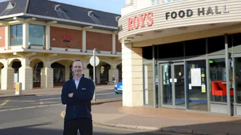 Paul Roy, wearing a navy Roys branded fleece and with his arms folded, stands in front of Roys Food Hall and Roys Department Store
