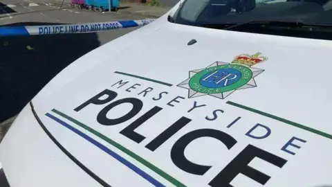 PA Media Close-up of a Merseyside Police vehicle's primarily white bonnet. Blue-and-white police cordon tape is behind the vehicle.