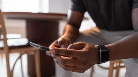 A man sits in a kitchen typing on his smartphone.