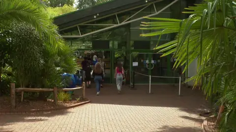 Paignton Zoo's entrance. A paved walkway goes into a glass structure with green metal frame. There are greens plant either side. 