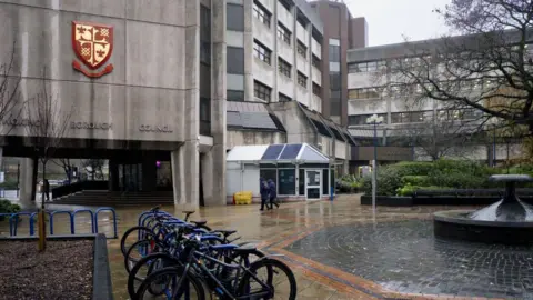 Getty Images A grey, brutalist building on a rainy day. A red and gold Woking Borough Council logo is fixed to the side of the building and there are four bicycles locked to a railing outside the building.