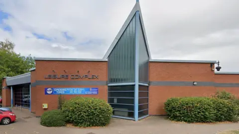 Google A single storey red brick building with a large vaulted glass wall in the middle. There is a sign that says Leisure Complex on the upper left side of the building and below it is a blue sign that says opening times