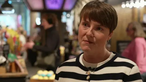 Kasey Swithenbank with short cropped brown hair and a black and white horizontal striped jumper standing in a Lush store with the till and colourful bath bombs behind her