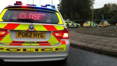 A Merseyside Police car flashing a 'Closed' sign in its back window with a row of police cars in the distance