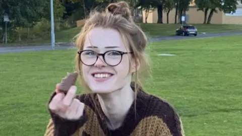Warrington Police / PA Brianna Ghey smiles at the camera while eating a chocolate. She has ginger hair tied in a bun and is wearing brown-rimmed glasses and a brown jumper.