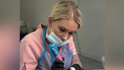 Belle Rose Lashes and Beauty Academy Kirsty Jackson is pictured using a tattoo gun. She wears a blue mask on her face, black gloves and pink uniform with a blue plastic overall over the top. Her blonde hair has been tied back.