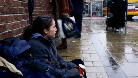 Getty Images A woman sitting on the pavement with her back against a red brick wall and a rucksack beside her. She is holding a disposable coffee cup. In the background there are people walking past and a bus stop with a bus at it.