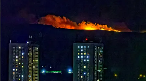 Alamy Orange flames on a dark hill with grey smoke billowing from them.
