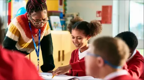 Getty Images A teacher with red and black braids placed in a bun is leaning over a table talking to pupils. A girl has two afro buns on the side of her head and is wearing a white polo shirt under a red cardigan. The teacher is wearing glasses and a red, orange, yellow, green and white wool jumper.