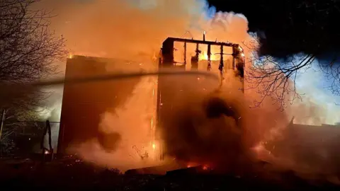 A part of the hotel, which can be seen badly damaged, is engulfed in smoke and a bright orange glow from the flames of the fire.