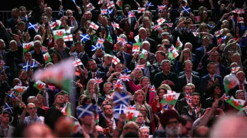 Getty Images People tightly packed into a stand wave small flags above their heads. There are blue and white saltires, Welsh red dragon flags and red and white St George's cross flags. 